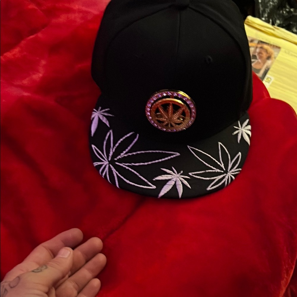 Black snap back hat w white pot leaf design on the bill and spinner style metal